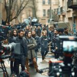 A bustling urban street filled with a large group of people, including crew members and equipment, suggesting a film production in progress.